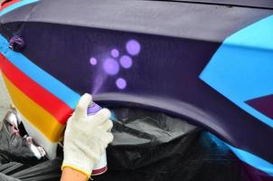 A hand with a spray can that draws a new colorful graffiti on the car. Photo of the process of drawing a graffiti on an automobile close-up. The concept of street art and illegal vandalism