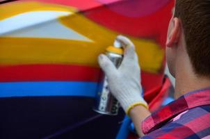 A young red-haired graffiti artist paints a new colorful graffiti on the car. Photo of the process of drawing a graffiti on a car close-up. The concept of street art and illegal vandalism