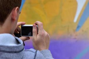 un joven grafitero fotografía su cuadro completo en la pared. el chico usa tecnología moderna para capturar un colorido dibujo de graffiti abstracto. centrarse en el dispositivo de fotografía foto