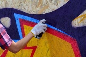 una mano con un bote de spray que dibuja un nuevo grafiti en la pared. foto del proceso de dibujar un graffiti en un primer plano de la pared de madera. el concepto de arte callejero y vandalismo ilegal