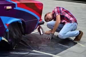 A young red-haired graffiti artist paints a new colorful graffiti on the car. Photo of the process of drawing a graffiti on a car close-up. The concept of street art and illegal vandalism