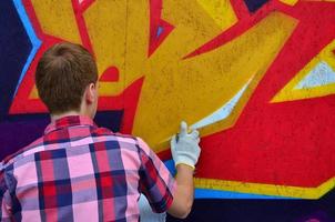 A young red-haired graffiti artist paints a new graffiti on the wall. Photo of the process of drawing a graffiti on a wall close-up. The concept of street art and illegal vandalism