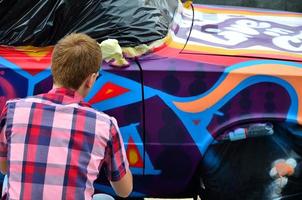 A young red-haired graffiti artist paints a new colorful graffiti on the car. Photo of the process of drawing a graffiti on a car close-up. The concept of street art and illegal vandalism