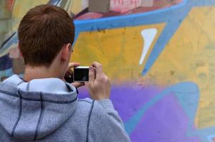 un joven grafitero fotografía su cuadro completo en la pared. el chico usa tecnología moderna para capturar un colorido dibujo de graffiti abstracto. centrarse en el dispositivo de fotografía foto