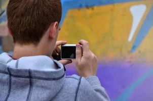 un joven grafitero fotografía su cuadro completo en la pared. el chico usa tecnología moderna para capturar un colorido dibujo de graffiti abstracto. centrarse en el dispositivo de fotografía foto