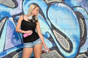 A young and beautiful sexy girl graffiti artist with a paint spray and gas mask on her neck stands on the wall background with a graffiti pattern in blue and purple tones photo