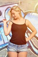 Chica rubia caucásica sexy en pantalones cortos de mezclilla y camiseta sin mangas negra posando contra la pared de graffiti durante el día al aire libre foto