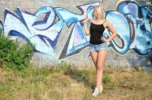 Chica rubia caucásica sexy en pantalones cortos de mezclilla y camiseta sin mangas negra posando contra la pared de graffiti durante el día al aire libre foto