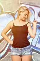 Sexy caucasian blonde girl in denim shorts and black tank top posing against graffiti wall in the daytime outdoors photo