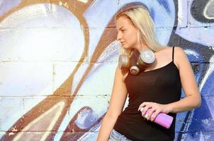 A young and beautiful sexy girl graffiti artist with a paint spray and gas mask on her neck stands on the wall background with a graffiti pattern in blue and purple tones photo