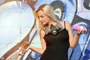 A smiling young and beautiful sexy girl graffiti artist with a paint spray and gas mask on her neck stands on the wall background with a graffiti pattern in blue and purple tones photo