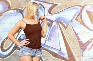 Chica rubia caucásica sexy en pantalones cortos de mezclilla y camiseta sin mangas negra posando contra la pared de graffiti durante el día al aire libre foto