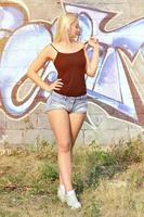 Sexy caucasian blonde girl in denim shorts and black tank top posing against graffiti wall in the daytime outdoors photo