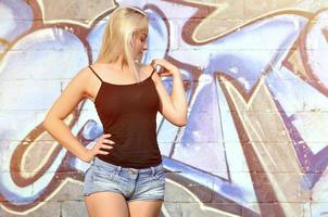 Chica rubia caucásica sexy en pantalones cortos de mezclilla y camiseta sin mangas negra posando contra la pared de graffiti durante el día al aire libre foto