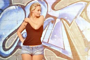 Sexy caucasian blonde girl in denim shorts and black tank top posing against graffiti wall in the daytime outdoors photo