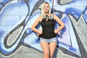 A smiling young and beautiful sexy girl graffiti artist with a paint spray and gas mask on her neck stands on the wall background with a graffiti pattern in blue and purple tones photo