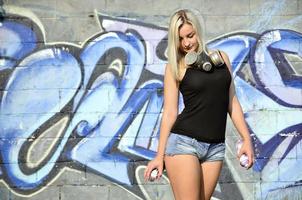 A young and beautiful sexy girl graffiti artist with a paint spray and gas mask on her neck stands on the wall background with a graffiti pattern in blue and purple tones photo