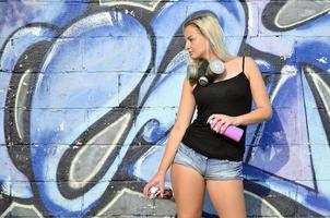 A young and beautiful sexy girl graffiti artist with a paint spray and gas mask on her neck stands on the wall background with a graffiti pattern in blue and purple tones photo