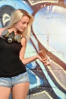 una joven y hermosa artista de graffiti sonriente y sexy con un spray de pintura y una máscara de gas en el cuello se encuentra en el fondo de la pared con un patrón de graffiti en tonos azules y morados foto