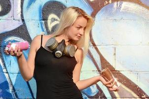 A smiling young and beautiful sexy girl graffiti artist with a paint spray and gas mask on her neck stands on the wall background with a graffiti pattern in blue and purple tones photo