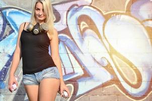 una joven y hermosa artista de graffiti sonriente y sexy con un spray de pintura y una máscara de gas en el cuello se encuentra en el fondo de la pared con un patrón de graffiti en tonos azules y morados foto