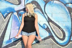 A young and beautiful sexy girl graffiti artist with a paint spray and gas mask on her neck stands on the wall background with a graffiti pattern in blue and purple tones photo