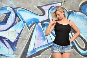 Chica rubia caucásica sexy en pantalones cortos de mezclilla y camiseta sin mangas negra posando contra la pared de graffiti durante el día al aire libre foto