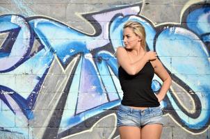 Chica rubia caucásica sexy en pantalones cortos de mezclilla y camiseta sin mangas negra posando contra la pared de graffiti durante el día al aire libre foto