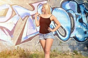 Chica rubia caucásica sexy en pantalones cortos de mezclilla y camiseta sin mangas negra posando contra la pared de graffiti durante el día al aire libre foto