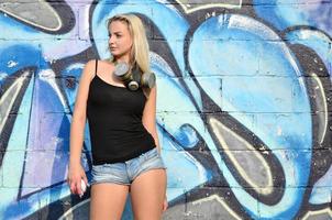 A young and beautiful sexy girl graffiti artist with a paint spray and gas mask on her neck stands on the wall background with a graffiti pattern in blue and purple tones photo