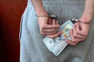 An arrested guy in gray pants with handcuffed hands holds a huge amount of dollar bills. Back view photo