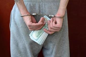 An arrested guy in gray pants with handcuffed hands holds a huge amount of dollar bills. Back view photo