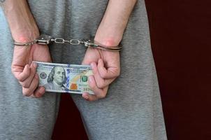 An arrested guy in gray pants with handcuffed hands holds a huge amount of dollar bills. Back view photo