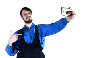 un joven barbudo con uniforme de trabajo se hace un selfie en un smartphone moderno, apuntándose a sí mismo con el dedo. un trabajador orgulloso se fotografía a sí mismo. aislado en blanco foto