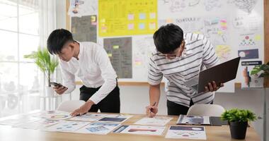 Two asian men checking to task on workplace desk while standing in office. One man holding laptop and writing on paper, One man using smartphone and writing notes on colorful sticky papers. video