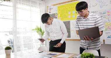 Two asian men checking to task on workplace desk while standing in office. One man holding laptop and writing on paper, One man using smartphone and writing notes on colorful sticky papers. video