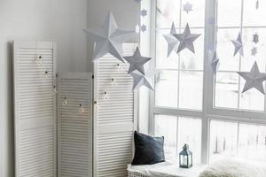Large bright window. On the window-sill is a gray knitted plaid with gifts and sparkles with herlands. Paper Christmas stars are suspended on the background of the window. photo