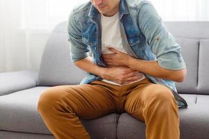 The man is sitting on a gray couch and holding his belly. Medicine and health concept, stomach problems. The man suffers from stomach ache, gastric problems. Abdominal pain, suffering and pain. photo