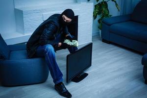 Robber man dressed in black hoodie stands with disguised face and holds a lot of money in his hands, stole a large amount, a thief man stole a TV photo