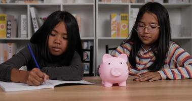 Two asian girls saving money with pink piggy bank while sitting at desk. Girl glasses putting coins into piggy bank and girl short hair writing on paper. Saving money concept. video