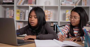 porträtt av två asiatisk studerande flickor Sammanträde på skrivbord på Hem. flicka kort hår och flicka glasögon inlärning uppkopplad via bärbar dator. ung kvinna skrivning en bok. utbildning begrepp. video