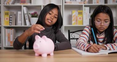 Zwei asiatische Mädchen sparen Geld mit rosa Sparschwein, während sie am Schreibtisch sitzen. Mädchen mit kurzen Haaren, die Münzen in ein Sparschwein stecken, und eine Mädchenbrille, die auf Papier schreibt. Geld sparen Konzept. video