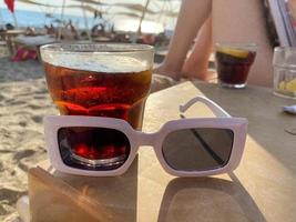 A glass with a soft drink brown cocktail with soda and whiskey and sunglasses on a table on the beach at the sea in a hotel on vacation in a paradise warm eastern tropical country resort photo