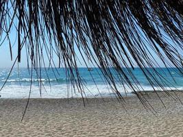 View of the blue sea with salt water through the yellow dry straw of the sun umbrella on the beach in the warm eastern tropical country southern paradise resort. The background photo