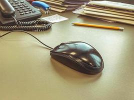 un ratón de computadora negro se encuentra en una mesa de oficina de madera. equipo de cómputo para el trabajo. mouse para clics, trabaje detrás del monitor. conveniencia del empleado foto