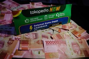 Jakarta, Indonesia in December 2022. A Tokopedia Now flyer above several hundred thousand rupiah notes scattered around. photo