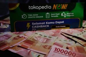 Jakarta, Indonesia in December 2022. A Tokopedia Now flyer above several hundred thousand rupiah notes scattered around. photo