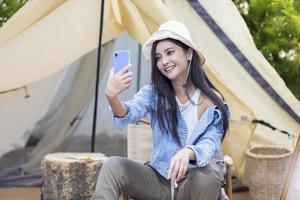 mujer asiática está usando un teléfono móvil para tomar una foto selfie en su tienda mientras acampa al aire libre durante el verano en el parque nacional para la aventura y el concepto de viaje activo