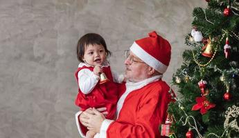 Santa Claus is lifting happy little toddler baby girl up and laughing cheerfully while helping to decorate christmas tree on the back for season celebration photo