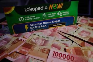 Jakarta, Indonesia in December 2022. A Tokopedia Now flyer above several hundred thousand rupiah notes scattered around. photo